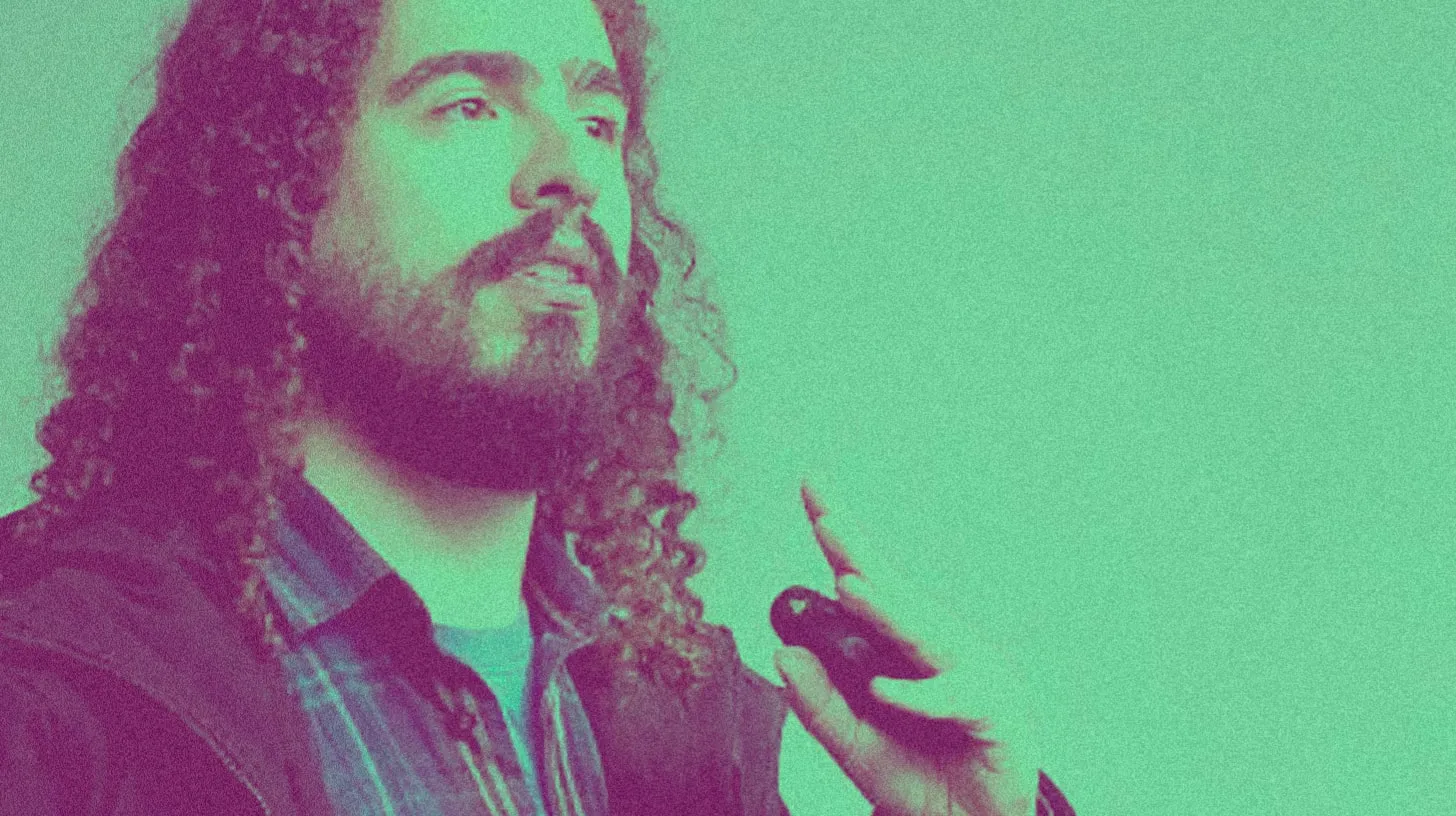 Retrato de homem de cabelos longos cacheados e barba, falando com um passador de slides na mão, em tons de verde e roxo